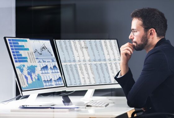 A businessman sitting at a desk, looking thoughtfully at two large flat screen monitors.