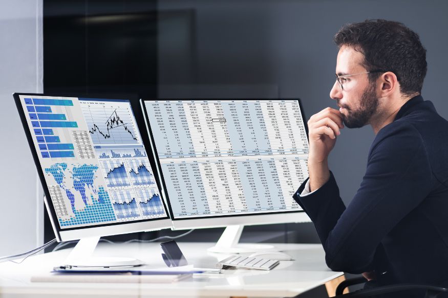 A businessman sitting at a desk, looking thoughtfully at two large flat screen monitors.