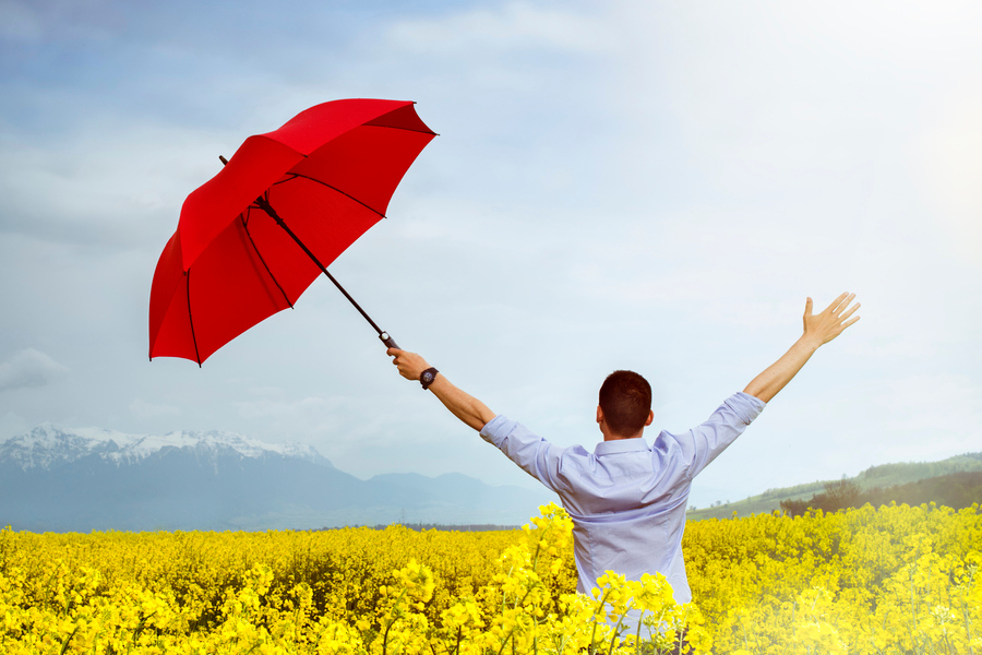 A man in a field of flowers, arms outstretched happily as they face away from the camera, an open red umbrella in their left hand.